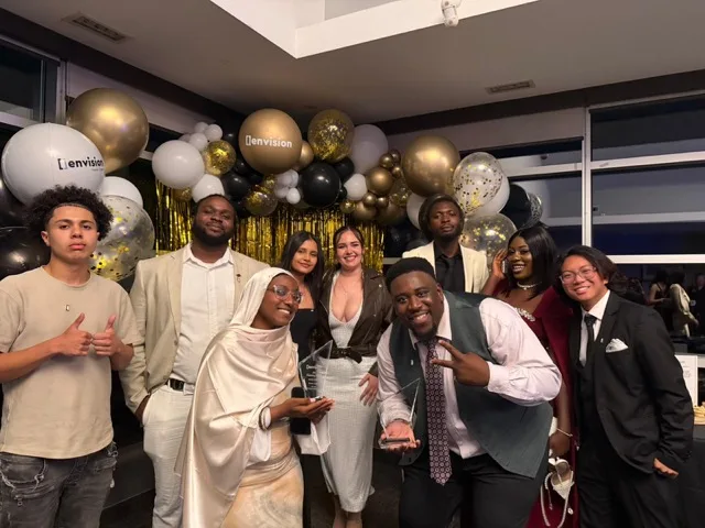The Legendz Consulting team at the Envision awards gala — nine team members in formalwear holding two crystal awards under a gold, black, and white balloon arch.